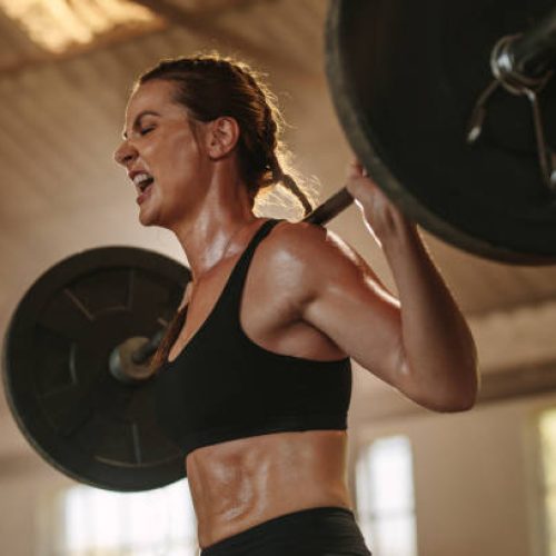 Female bodybuilder doing exercise with heavy weight bar. Fitness woman sweating from squats workout at gym. Female putting effort and screaming while exercising with heavy weights.